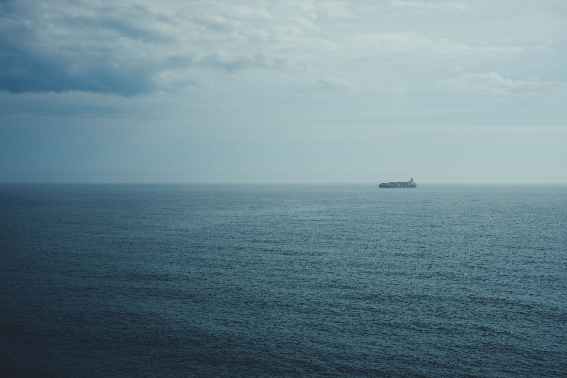 A distant ship navigates the open ocean beneath a cloudy sky